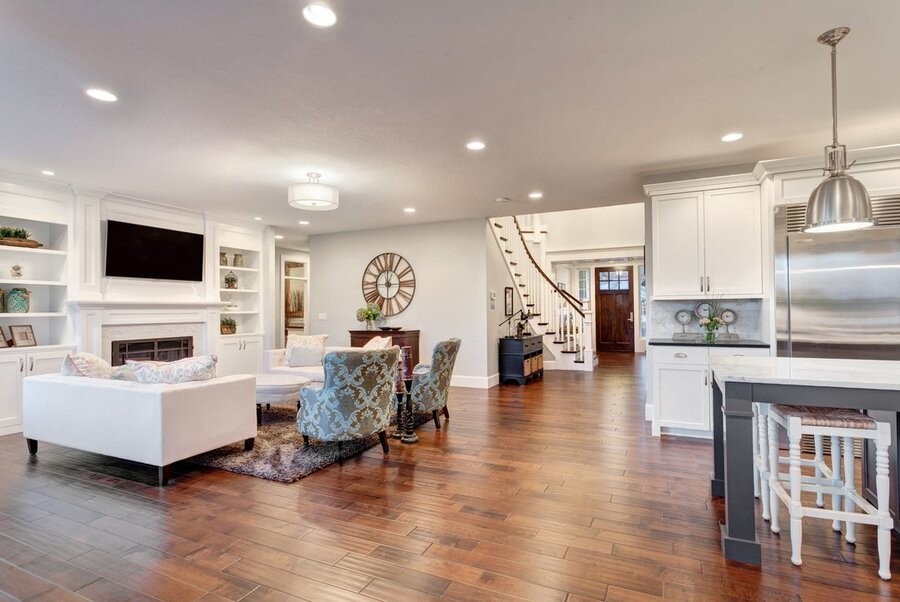An open living space connecting a living room to kitchen space with a foyer and staircase in the background, all illuminated by smart lighting fixtures.