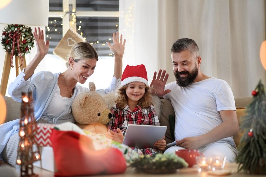 Two parents and a young daughter on a couch waving at a laptop with holiday decor surrounding them.